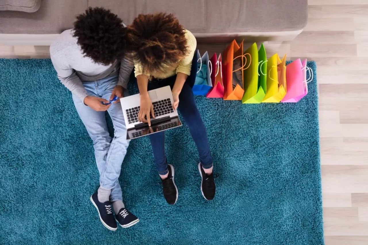 Young Couple Shopping Online On Laptop With Multi Colored Shopping Bags On Sofa